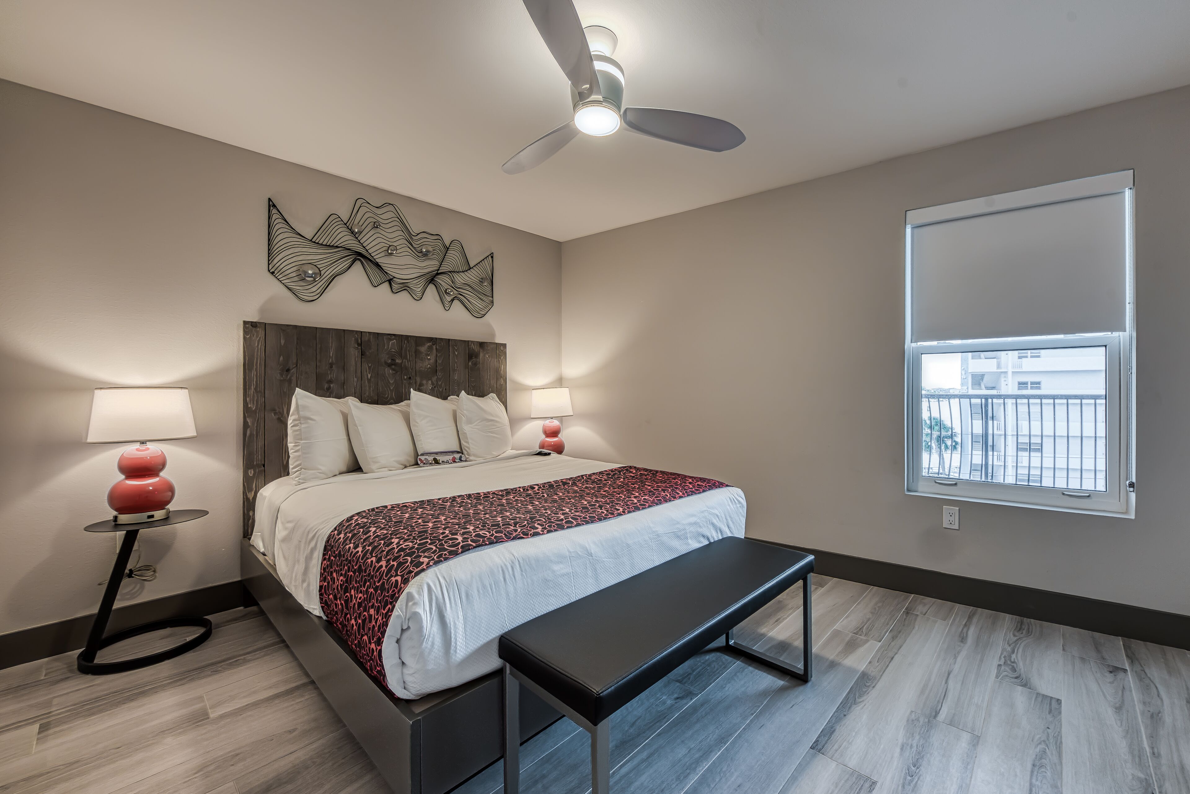 A bedroom with a bed, bedside lamps, a ceiling fan, a bench, and a window with a view.
