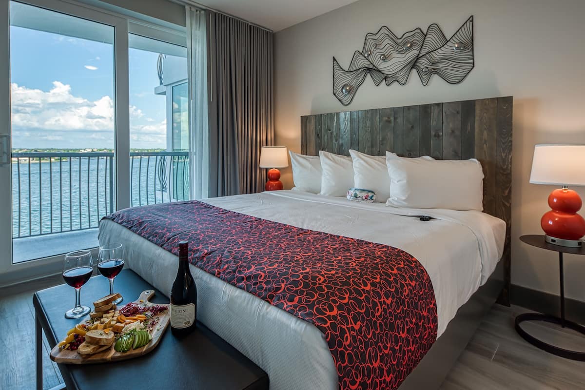 Une chambre d’hôtel avec un grand lit, une vue sur la mer, une tête de lit en bois et une table avec du vin et des amuse-gueules.