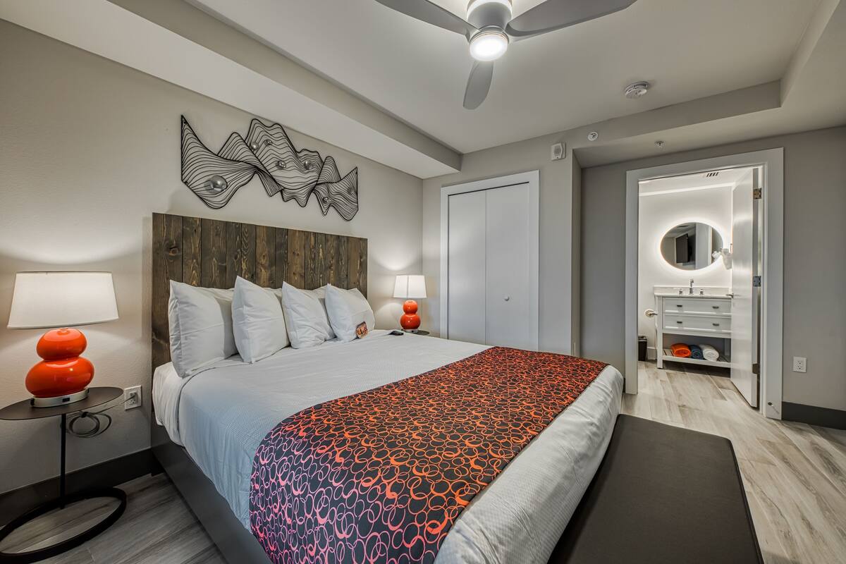 Une chambre moderne comprenant un lit, deux tables de chevet avec des lampes, un ventilateur de plafond, une tête de lit en bois, un grand tableau décoratif mural, un dressing et un miroir.