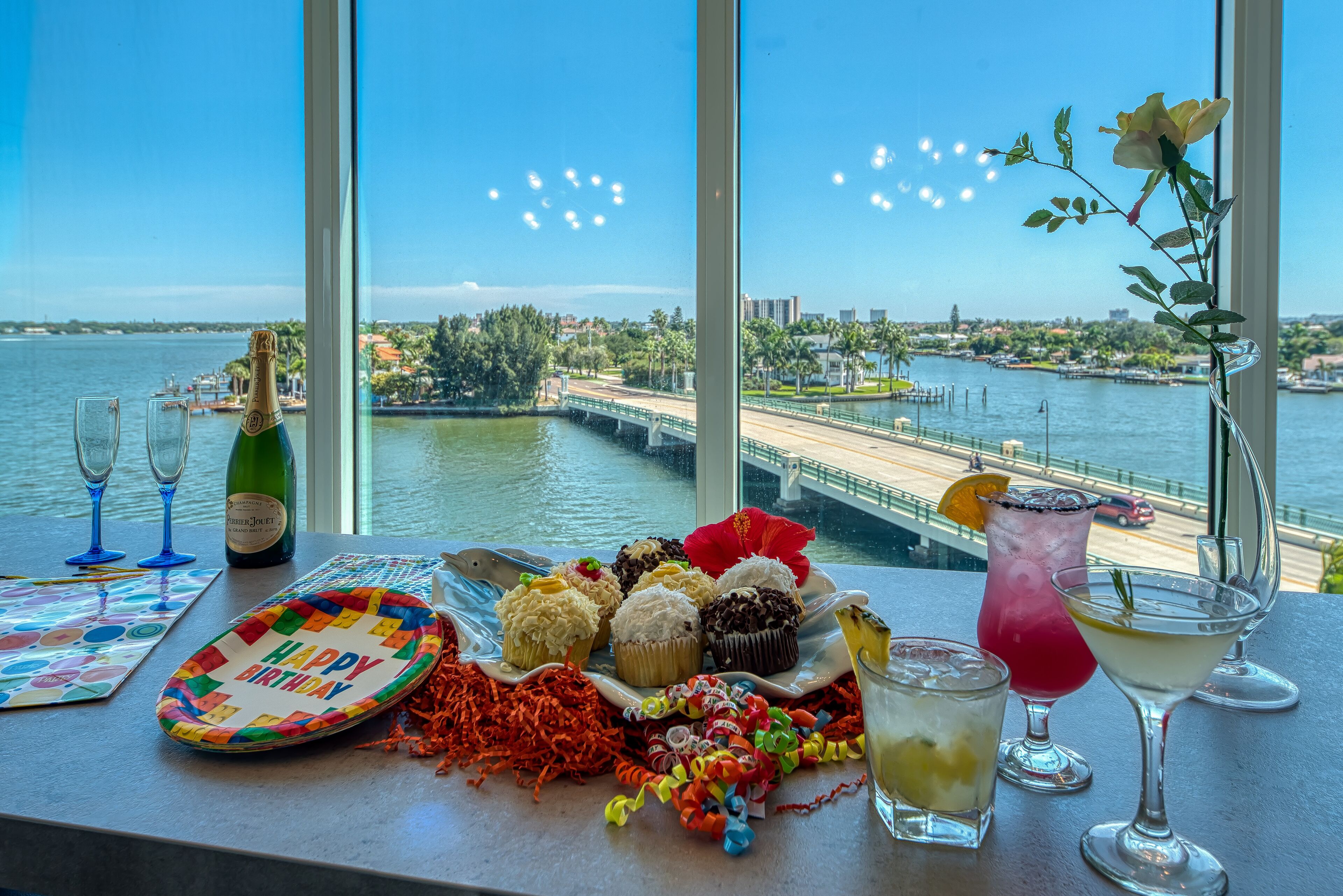 Une table avec une bouteille de champagne, une assiette d’anniversaire aux couleurs vives, des cupcakes et des boissons, donnant sur une rivière avec un pont et des bâtiments.