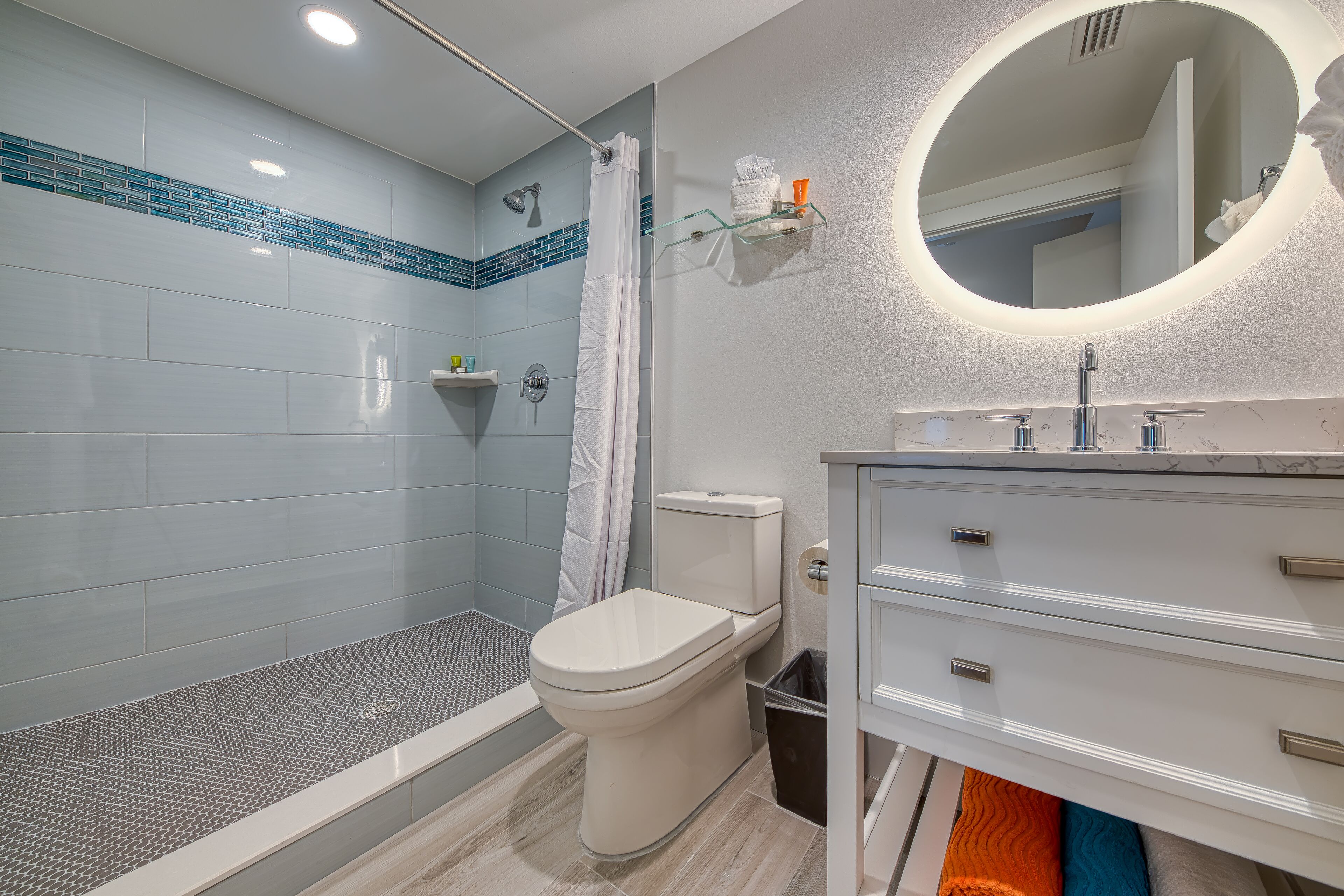 A modern bathroom with a glass shower enclosure, a toilet, a vanity with a mirror, and a small shelf with toiletries.