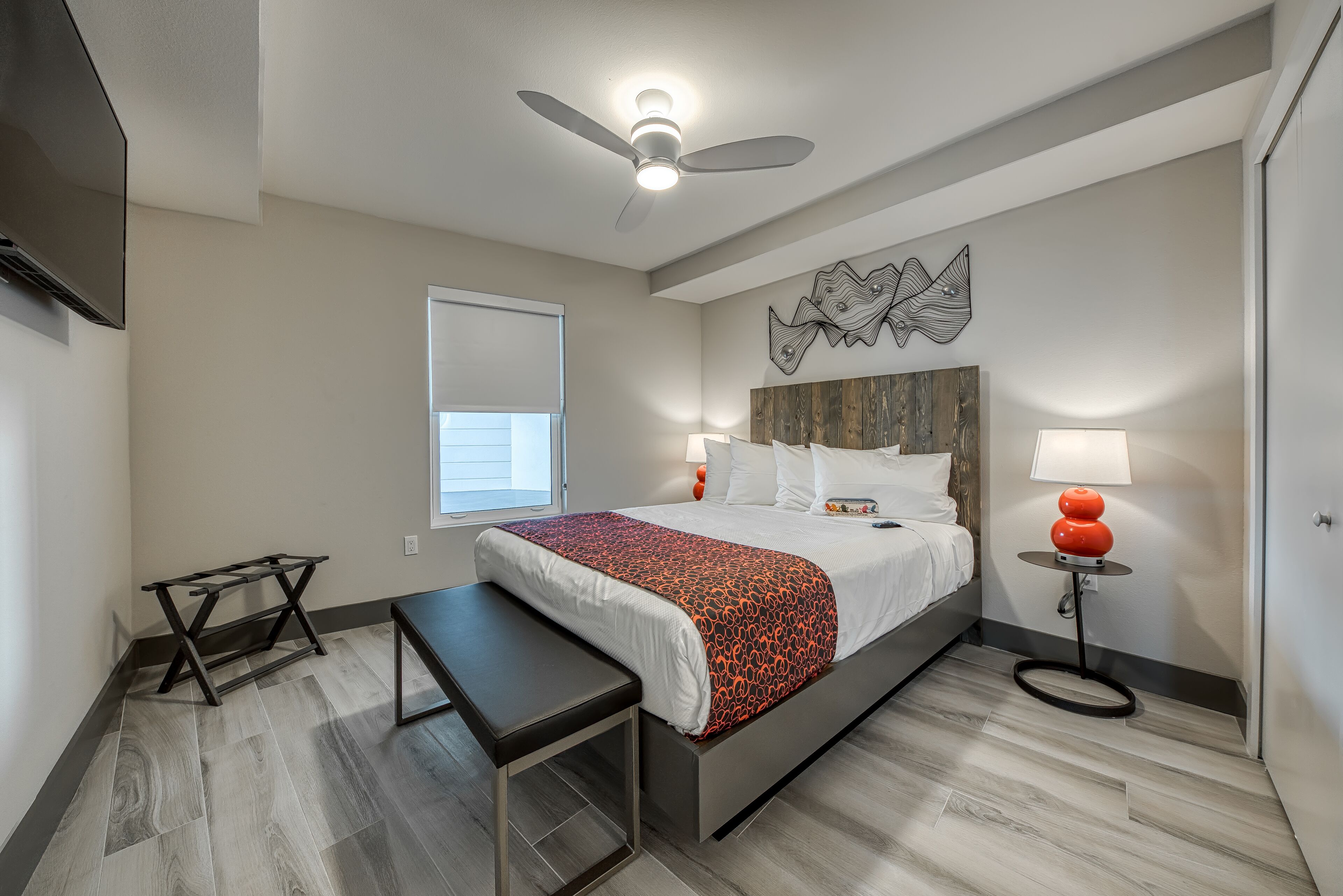 Una habitación de hotel moderna con una cama, un escritorio, un banco, un ventilador de techo, una obra de arte en la pared y una mesita de noche con una lámpara.
