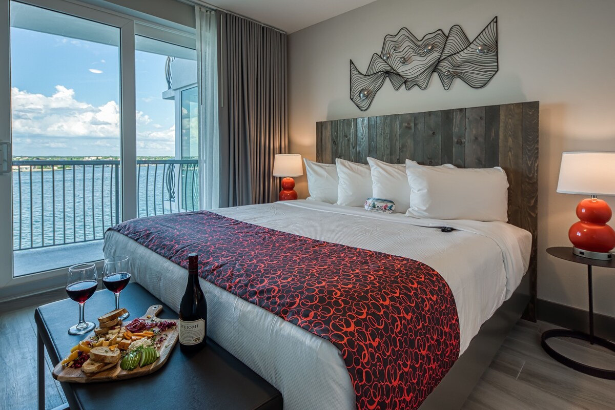 A hotel room with a large bed, a view of the sea, a wooden headboard, and a table with wine and snacks.