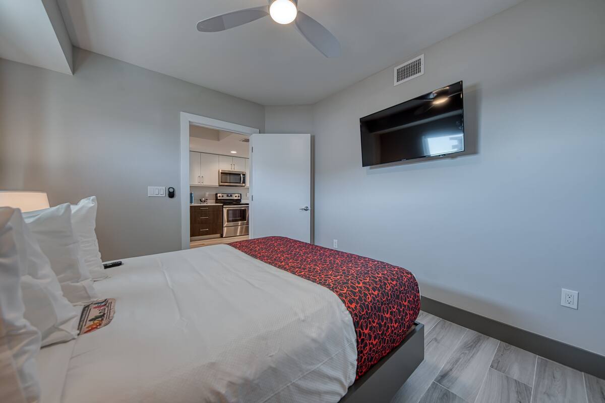 Une chambre à coucher avec un lit, un ventilateur de plafond, un téléviseur fixé au mur et, au fond, une petite cuisine.