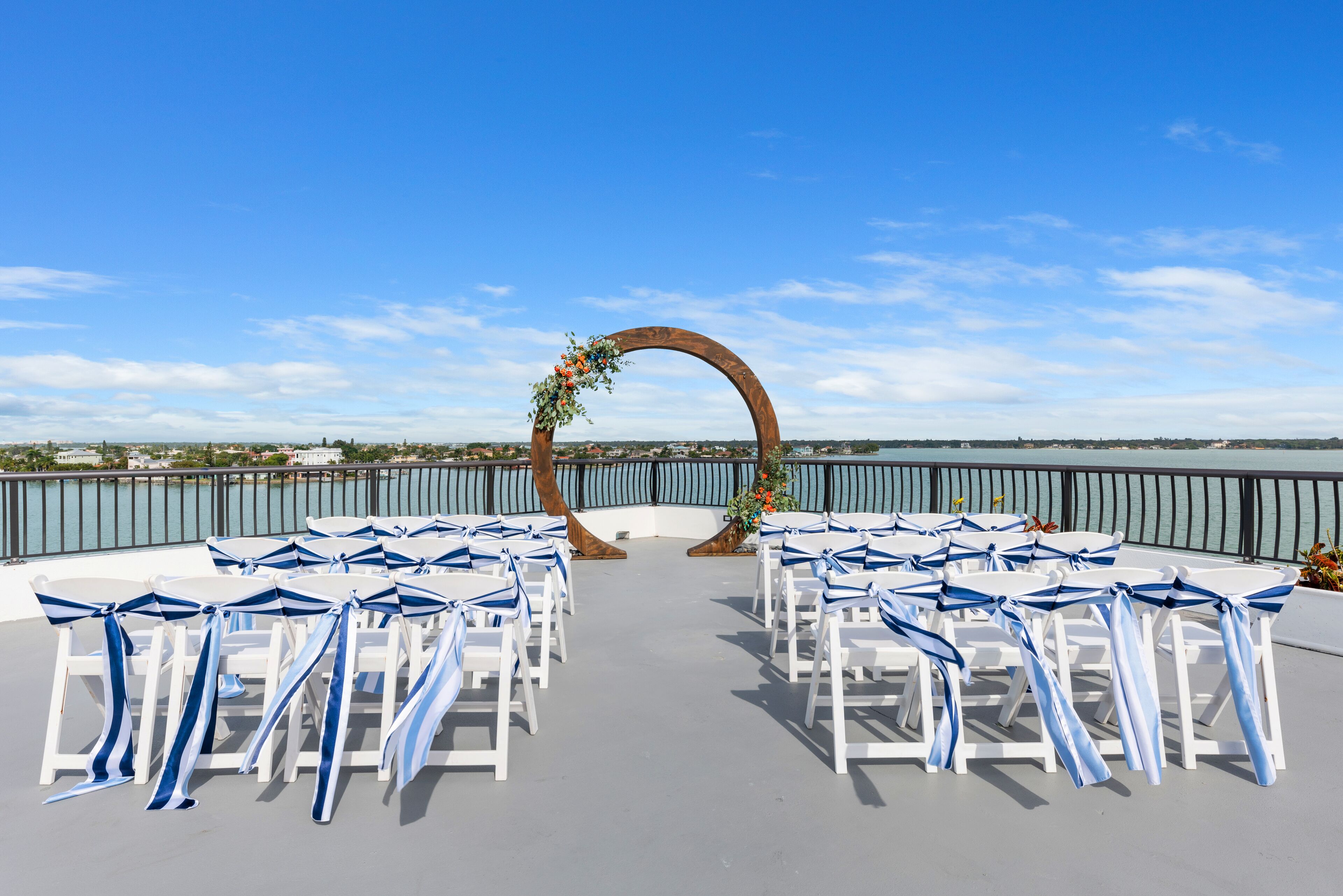 An outdoor seating area with white tables and chairs, arranged in rows facing a decorative archway with greenery and flowers.