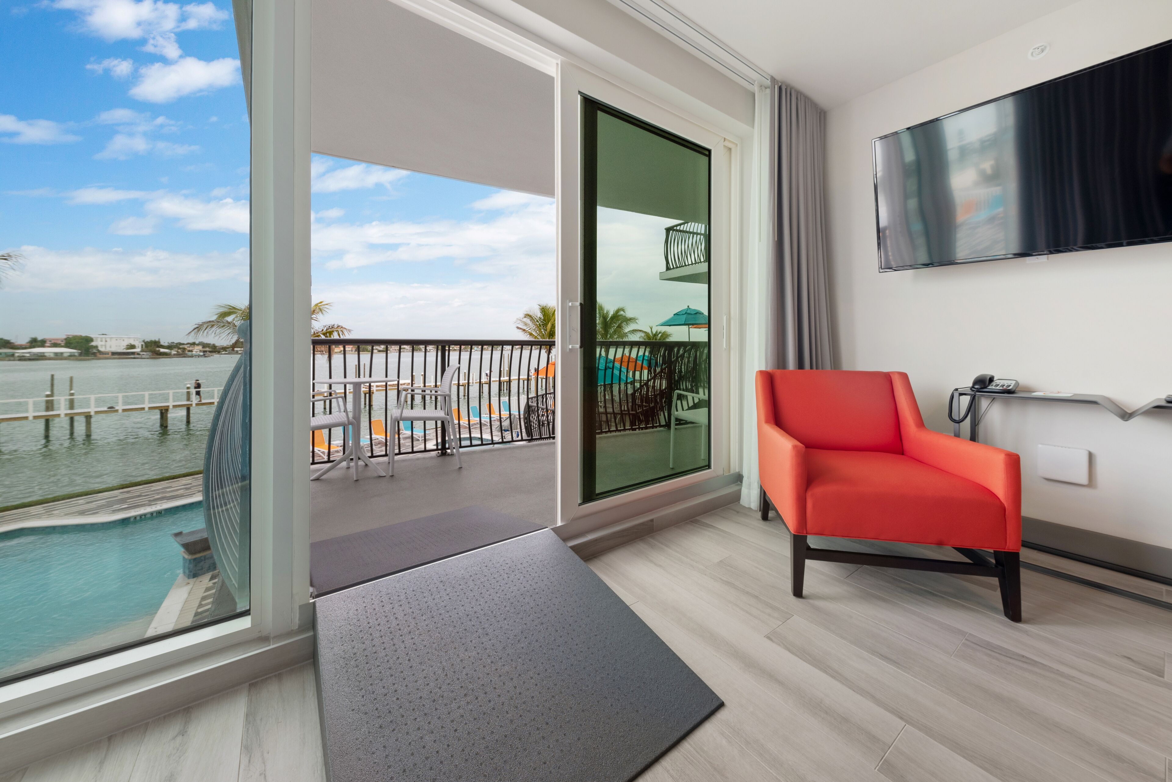 A modern hotel room with a large sliding glass door leading to a balcony with a pool, a flat-screen TV mounted on the wall, and a bright orange chair.