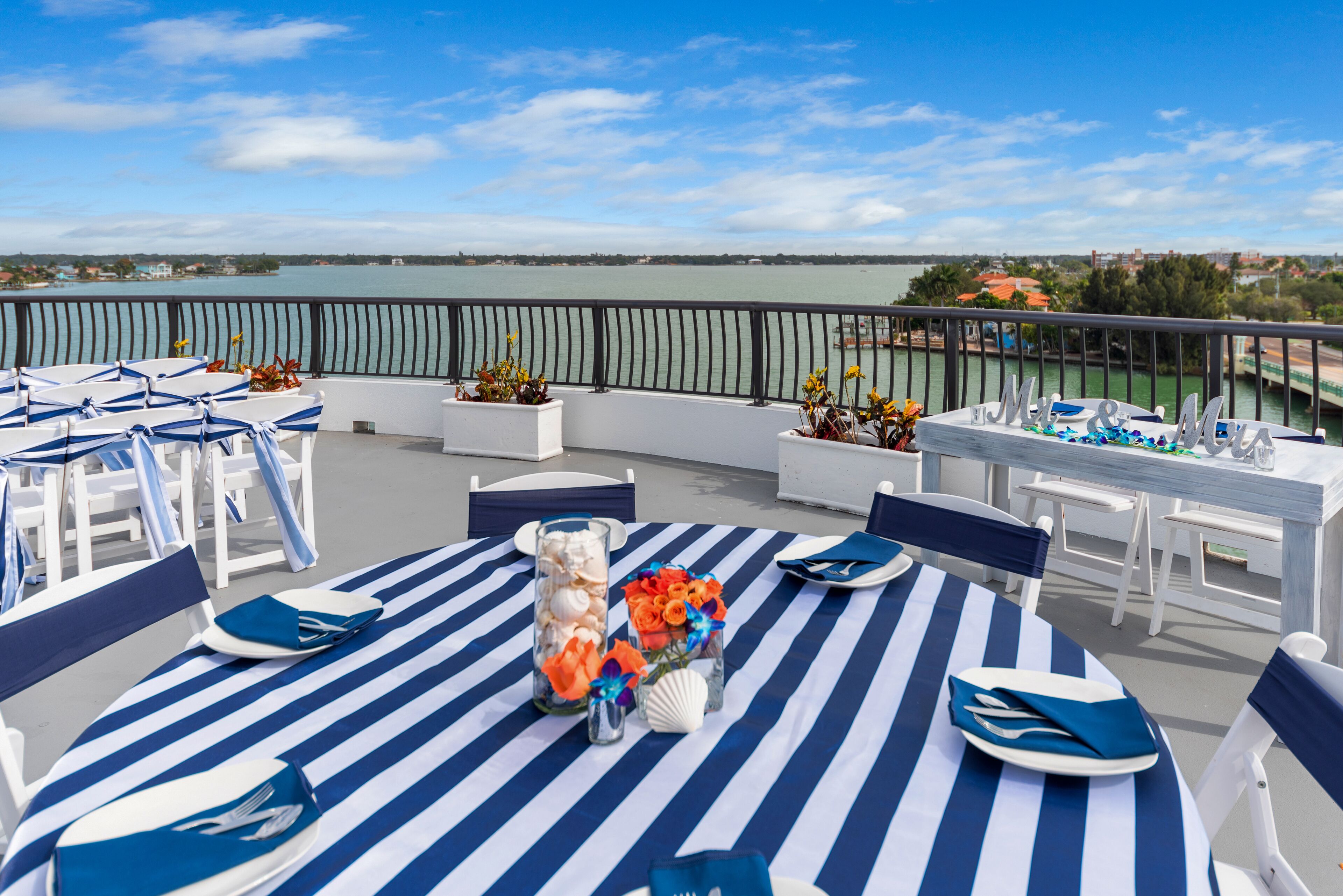 A terrace with a view of the water, featuring striped tablecloths, white chairs, and a table set with plates and napkins.