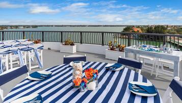 A terrace with a view of the water, featuring striped tablecloths, white chairs, and a table set with plates and napkins.
