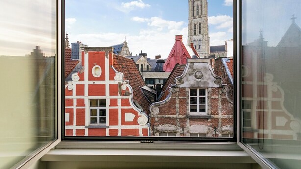 A window view of a cityscape with historic buildings, including a prominent church tower. - Lord of Ghent Luxurious apartment in historical center of Ghent (Gent)