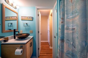 A bathroom with a large mirror, a bowl sink, and a toilet. There are two framed artworks on the wall and a curtain with a nautical theme.