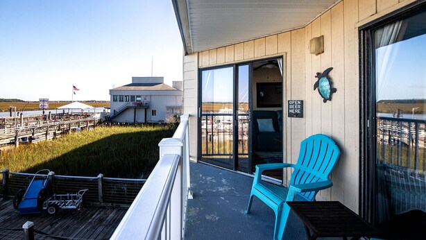 A deck with a blue chair and a cart, a building with a sign reading "OPEN BEER HERE", and a view of a marina with boats and a flag.