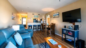A modern living room with a blue sofa, a coffee table, and a TV. There is a kitchen area with a bar and stools.