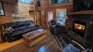 A log cabin interior with a fireplace, leather sofa, armchair, and a large window with a view of the outdoors. - Cozy Log Home, Centrally Located, 2 Master Suites, Hot Tub, Game and Movie Rooms (Sevierville)