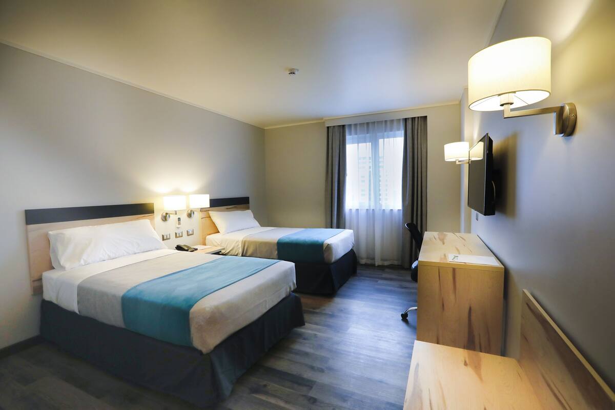 Ein Hotelzimmer mit zwei Betten, einem Schreibtisch, einem Flachbildfernseher und einem Fenster mit Vorhängen.