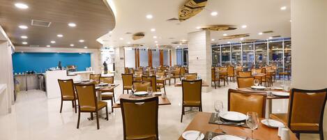 A spacious restaurant with multiple dining tables and chairs, a blue accent wall, and a modern ceiling design.