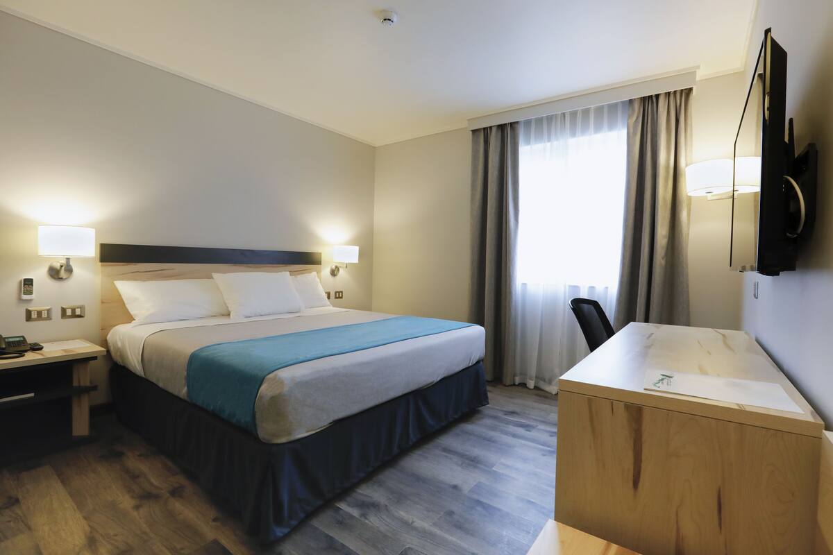 Ein Hotelzimmer mit Bett, Nachttischen, Schreibtisch, Sessel, Fernseher und einem Fenster mit Vorhängen.
