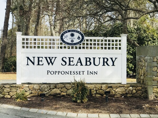 A sign for New Seabury Popponset Inn, featuring a logo with a sun, a bird, and a wave, and a white picket fence.