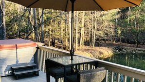 Une terrasse en bois avec une table et des chaises, un grand parasol et une vue sur un lac entouré d’arbres.