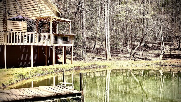 Une cabane en bois avec un quai s’avançant dans un étang paisible, le tout entouré d’arbres.