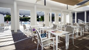 A covered outdoor area with white tables and chairs, a pool in the background, and a view of a building.