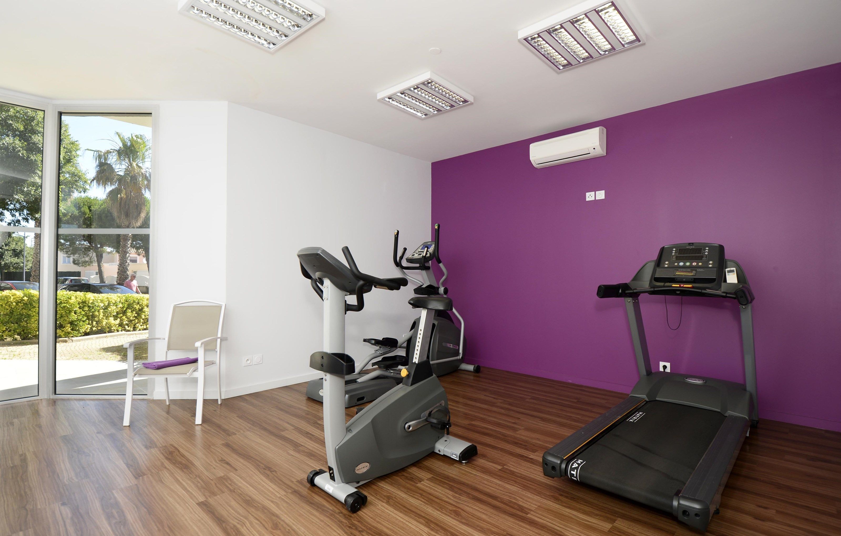 A fitness room with a treadmill, elliptical, and chair, large windows, and a purple wall.