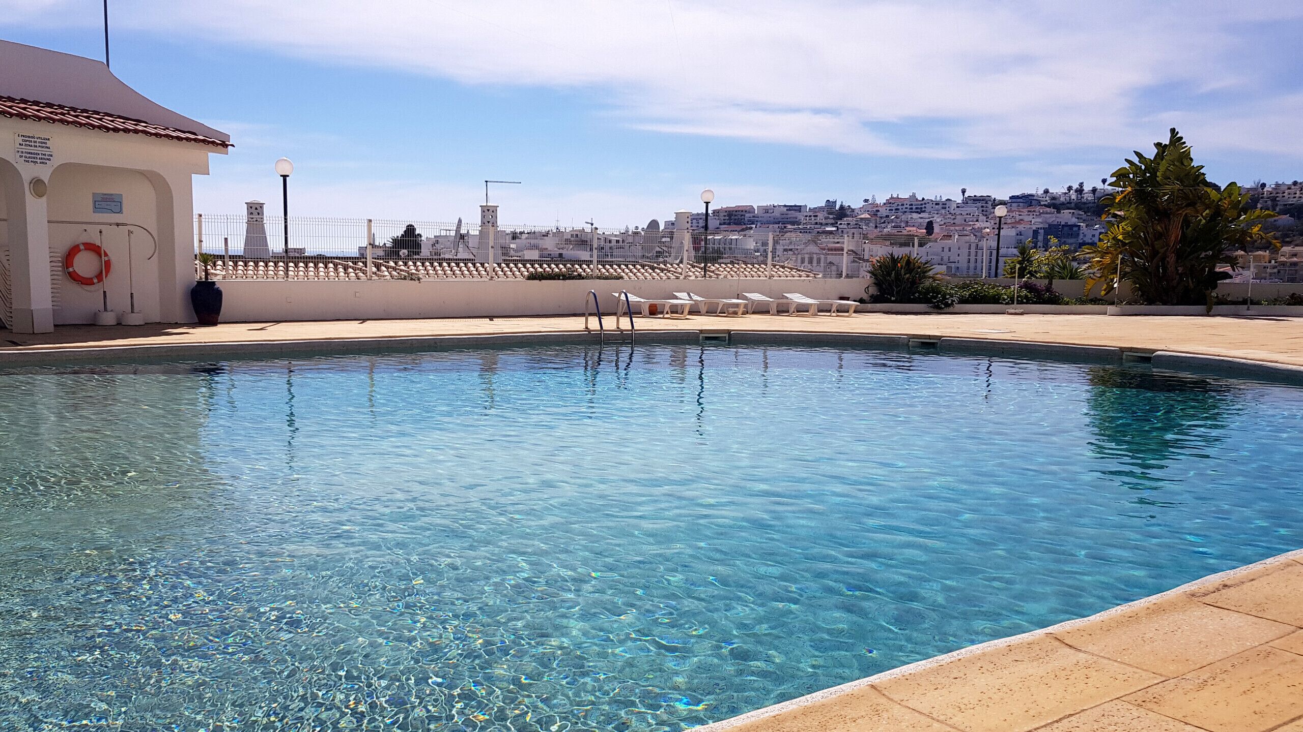 Una piscina cristalina con un salvavidas, una valla y una vista del paisaje urbano al fondo.