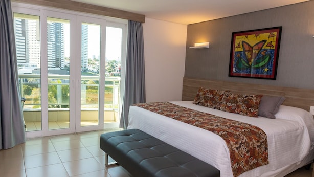 Une chambre d’hôtel avec un grand lit, un banc, un balcon avec des rideaux et un tableau coloré au mur.