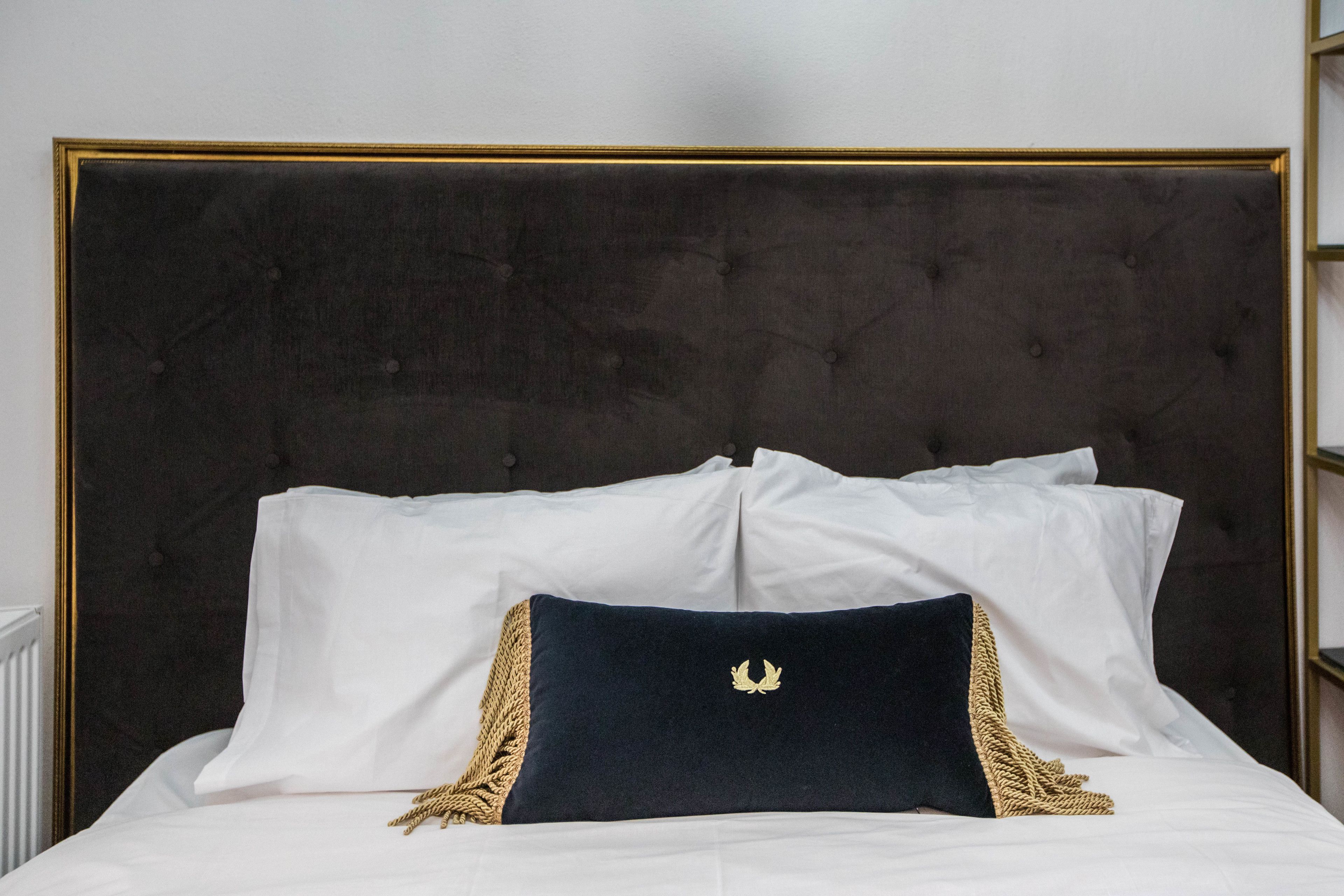 Una cama con cabecera oscura, almohadas blancas y una almohada oscura con un emblema dorado.