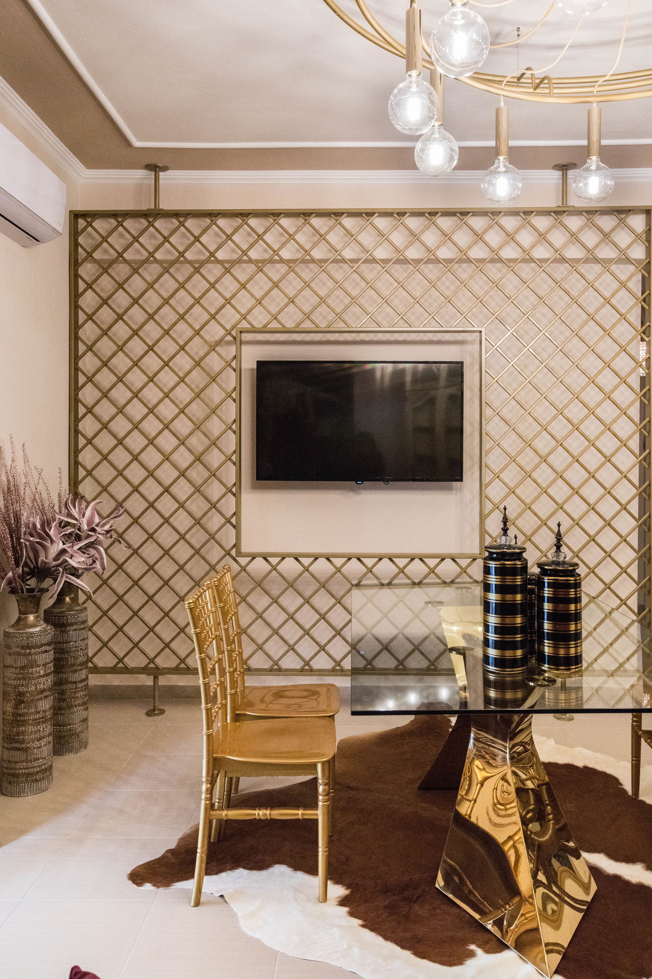 Een moderne eetruimte met een glazen tafel, gouden stoelen, een wandgemonteerde televisie en decoratieve vazen.