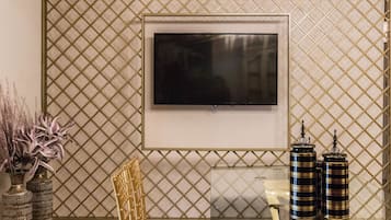 Een moderne eetruimte met een glazen tafel, gouden stoelen, een wandgemonteerde televisie en decoratieve vazen.