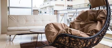 A wicker chair with a cushion, a sofa, and a small round table on a balcony with a view of buildings.