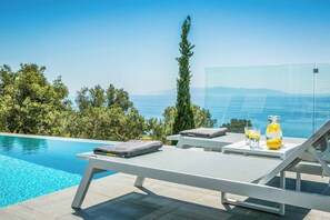 Area piscina con lettini, un tavolo con bevande e vista libera sul mare e sulle montagne.