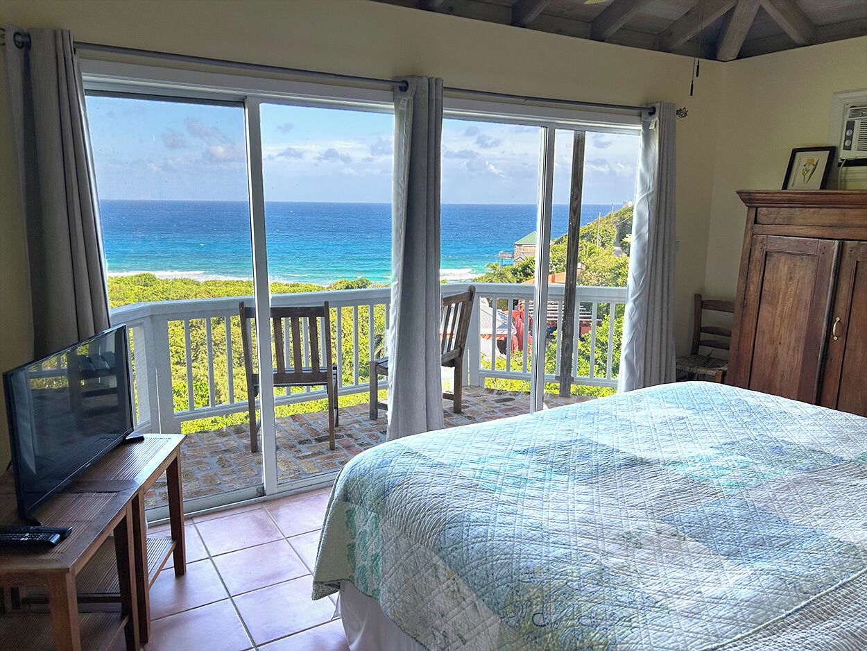 A bedroom with a large bed, a TV on a wooden table, and a balcony with a view of the ocean.