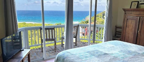 A bedroom with a large bed, a TV on a wooden table, and a balcony with a view of the ocean.