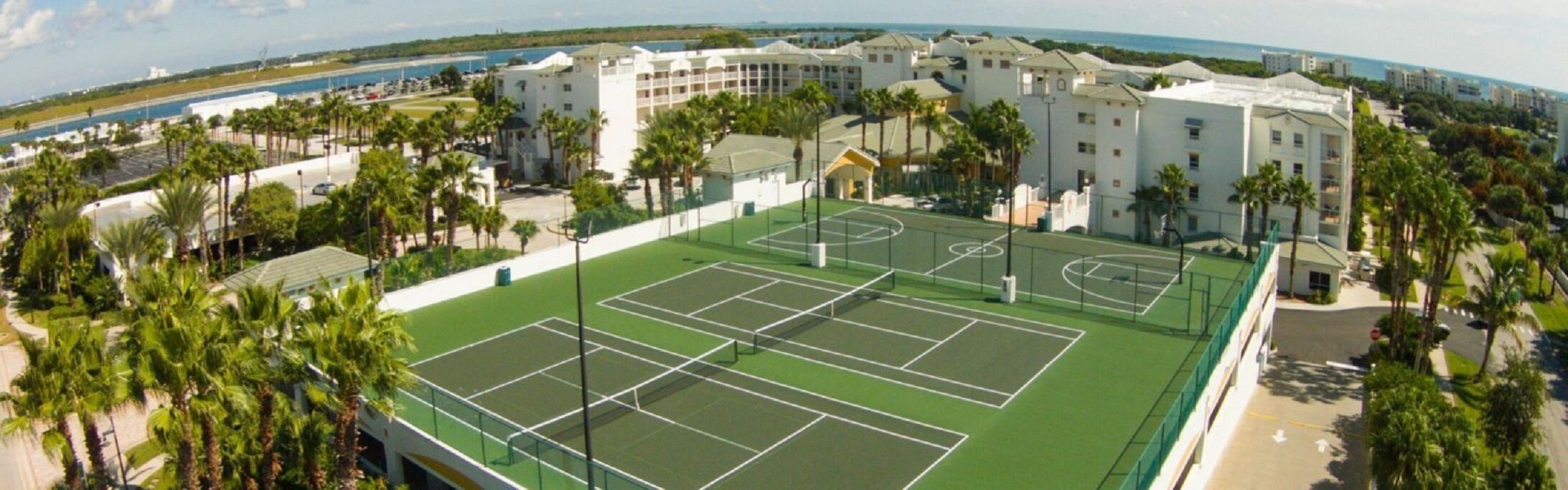 A tennis court surrounded by palm trees and a building with multiple floors.