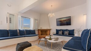 A modern living room with a fireplace, seating areas, and a view of the outdoors. - Rodanthi Hospitality (Faistos)
