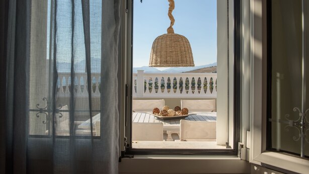 A view through an open window with a wicker hanging light fixture, a balcony with a table and chairs, and a decorative railing. - Rodanthi Hospitality (Faistos)