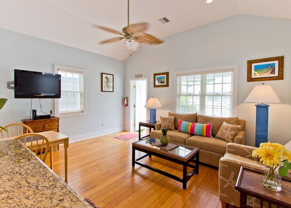 A spacious living room with hardwood floors, a ceiling fan, a flat-screen TV, a sofa with colorful pillows, a coffee table, and a dining area with a wooden table and chairs.