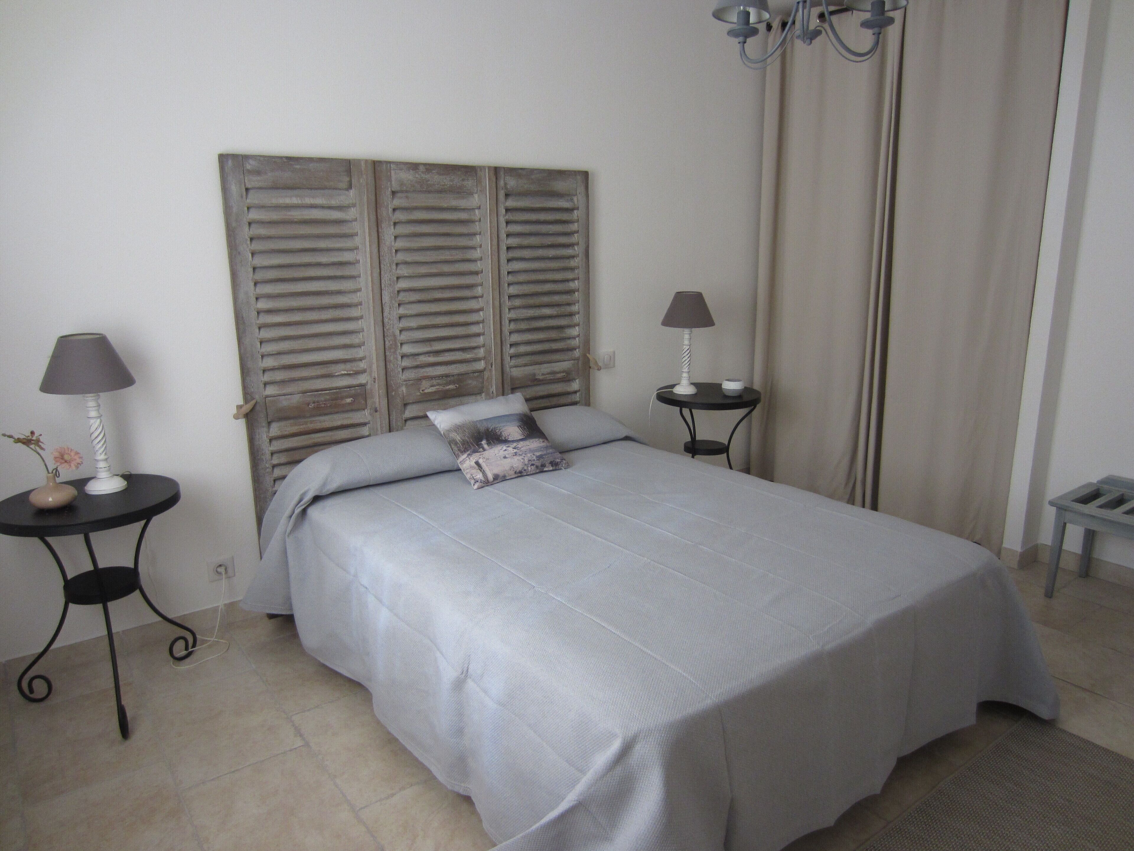 A neatly made bed with a grey bedspread and pillows, two bedside tables with lamps, a wooden headboard, and a chandelier.
