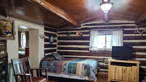 A log cabin room with a bed, a wooden chair, a television on a wooden stand, and a window with curtains. - Historic Moose Lodge Cabin (South Fork)