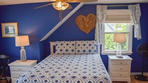 Una camera da letto con una coperta a motivi, una ventola a soffitto, una decorazione a forma di cuore, una finestra con tende e una lampada da comodino.