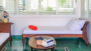 Una habitación con cama, una mesita con libros, un banco y una ventana con persianas.