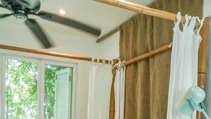 Un dormitorio con cama, ventilador de techo, ventana con cortinas y mesita de noche con libros.
