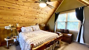 A wooden cabin bedroom with a bed, a ceiling fan, a window with curtains, and a lamp. - Mtn Views/Large Master Suite/Hot Tub/Wi-Fi (Bryson City)