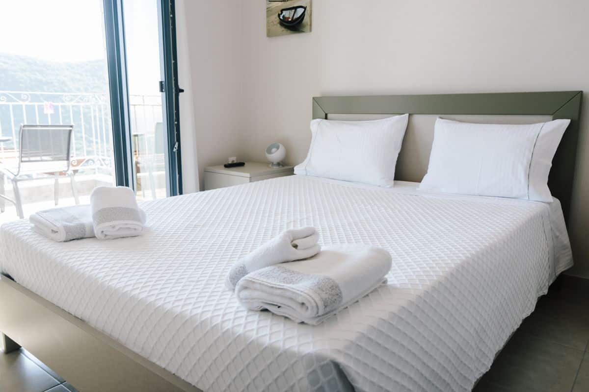 A neatly made bed with white bedding and two pillows, a bedside table with a lamp, and a view of a balcony with a chair.