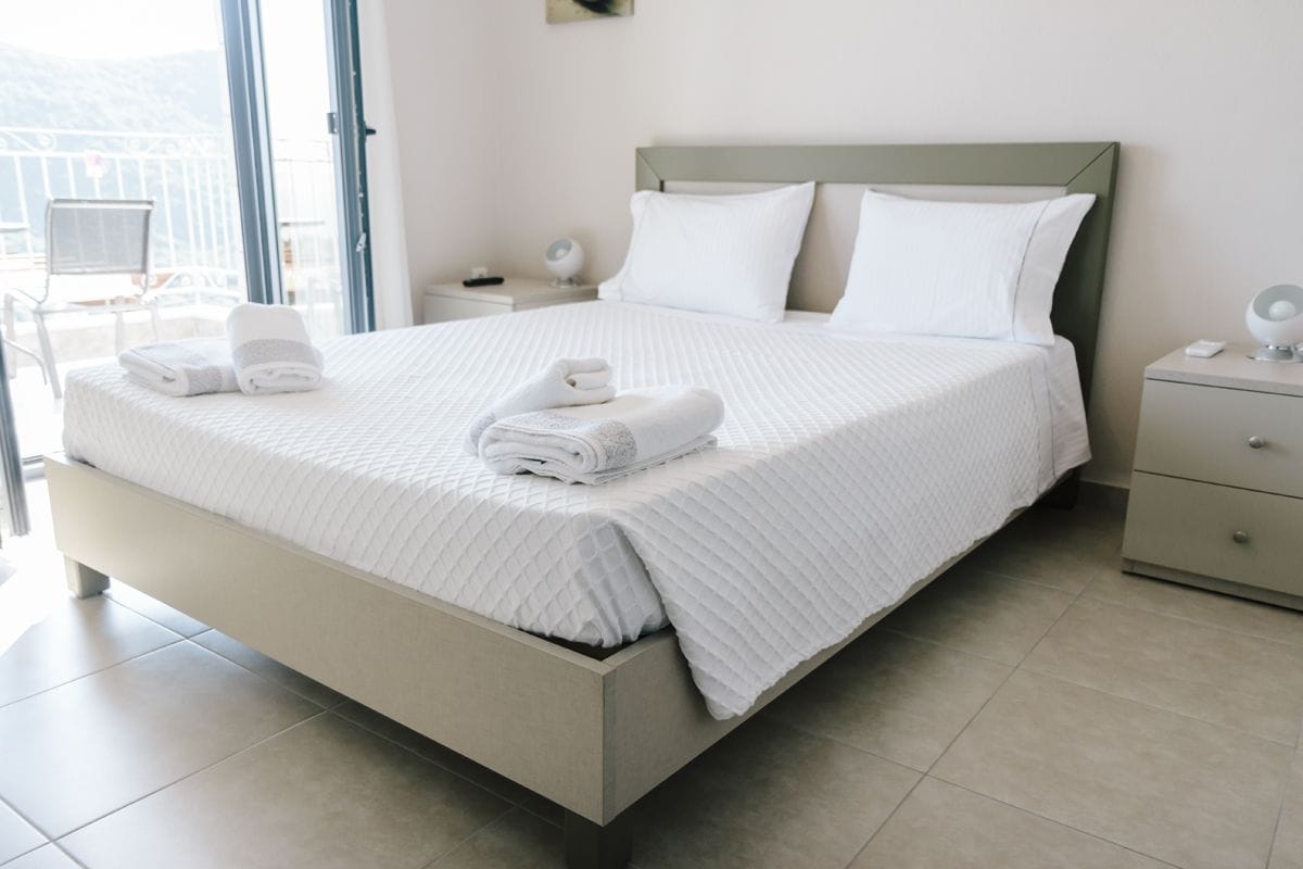 Cama bem arrumada com lençóis e almofadas brancas, mesa de cabeceira com abajur e vista para o exterior através de uma grande janela.