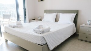 A neatly made bed with white linens and pillows, a bedside table with a lamp, and a view of the outdoors through a large window. - Villa Mons & Mare (Igoumenitsa)