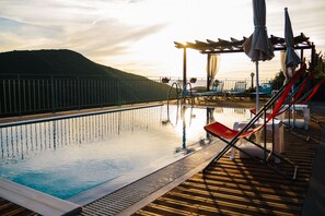 Zona de piscina con sillas de descanso y sombrillas, rodeada por una terraza de madera y una barandilla de metal.