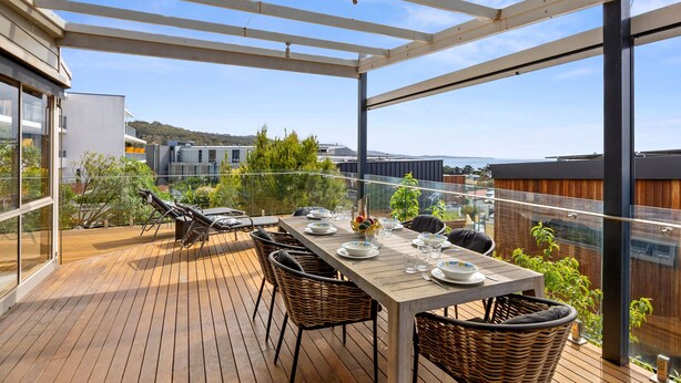 An outdoor patio with a wooden table set for dining, wicker chairs, and a glass railing overlooking a cityscape. - Boston Beach House (Lorne)