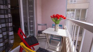 A balcony with a white table, red and yellow chairs, a vase with red flowers, and a view of a building with balconies.